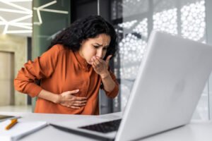 Sick business woman working at work, having severe nausea and abdominal pain, latin american working inside office building using laptop